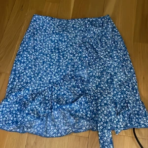 Blå blommig omlottkjol - Charmig blå omlottkjol med ett vitt blommönster. Kjolen har volangdetaljer och knytband i midjan för en justerbar passform. Perfekt för en somrig look! Man kan knyta rosett 