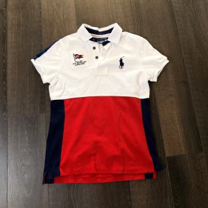 Ralph lauren pike - Snygg pikétröja från Ralph Lauren i vitt och rött med marinblå detaljer. Tröjan har korta ärmar och en klassisk krage med knappar. Den ikoniska loggan är broderad på bröstet. Perfekt för en stilren look! Mycket bra skick, går ej och få tag på!