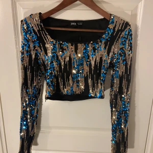 Paljettopp  - Säljer en glittrig svart topp från Shein med blå och guldiga paljetter. Toppen har axelvaddar och dragkedja på sidan. 