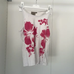 Vit linnetopp med rosa blommönster från Tribute - Säljer en vit linnetopp från Tribute med ett snyggt rosa blommönster på framsidan. Perfekt för varma dagar med sin ärmlösa design och bekväma passform. 🌸