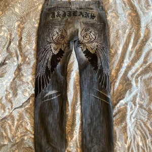 Unika jeans med vingbroderi från J&J Jeans - Säljer ett par asballa jeans från J&J Jeans. De är straight leg, blåa med ett unikt broderi av vingar och mönster på baksidan. Inga defekter, knappen är inte originell utan påsydd. Skulle nog säga att midjemåttet är emellan 40-42, OBS ej stretchiga. 