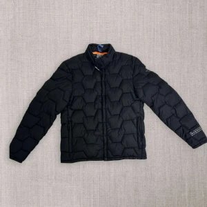 Svart quiltad jacka från Replay - Säljer en snygg svart quiltad jacka från Replay. Den är vattenavvisande och perfekt för resor med sin mångsidiga design. Jackan har en cool hexagonmönstrad quiltning och långa ärmar. Insidan är i en kontrasterande orange färg som ger en extra touch. Perfekt för höst och vår!