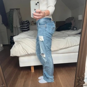 Blå jeans med slitningar - Säljer ett par blå jeans med coola slitningar på låren och knäna. De har en klassisk femficksdesign och en knappgylf. Perfekta för en avslappnad look.