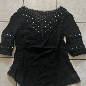 Svart blus med spets och pärlor - Elegant svart blus med trekvartsärm och vackra spetsdetaljer. Blusen har pärlor på spetsen vid halsringningen och ärmarna, vilket ger en lyxig känsla. Perfekt för en stilfull look.