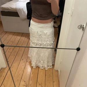 Vit långkjol med spetsdetaljer - Charmig vit långkjol med vackra spetsdetaljer och broderade blommor. Kjolen har en luftig och romantisk stil, perfekt för varma dagar. Den är tillverkad i ett lätt och bekvämt material som ger en skön passform.