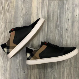 Burberry Sneakers - Snygga äkta svarta sneakers med ett klassiskt rutigt mönster i beige och brunt på sidorna. Skorna har en vit sula och svarta snören med röda detaljer vid snörningen. Perfekta för en stilren look.