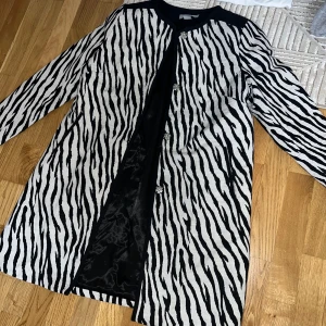 Zebramönstrad kappa med knappar - Snygg zebramönstrad kappa med knappar framtill. Den har långa ärmar och en stilren design som passar perfekt för att sticka ut. Kappan är i svart och vit med en elegant knäppning.