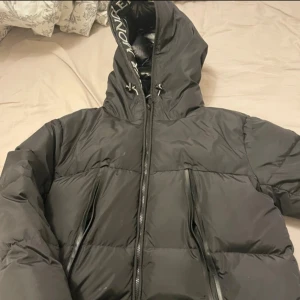 Svart dunjacka från Moncler - Snygg svart dunjacka från Moncler med huva och dragkedja. Jackan har Moncler-logga på ärmen och insidan av huvan. Perfekt för kyliga dagar.