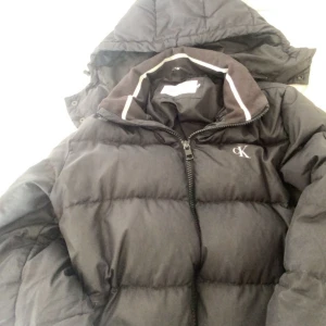 Svart dunjacka från Calvin Klein Jeans - Snygg svart dunjacka från Calvin Klein Jeans med huva och dragkedja. Perfekt för kyliga dagar med sin stilrena design och varma fyllning. Jackan har en liten CK-logga på bröstet.