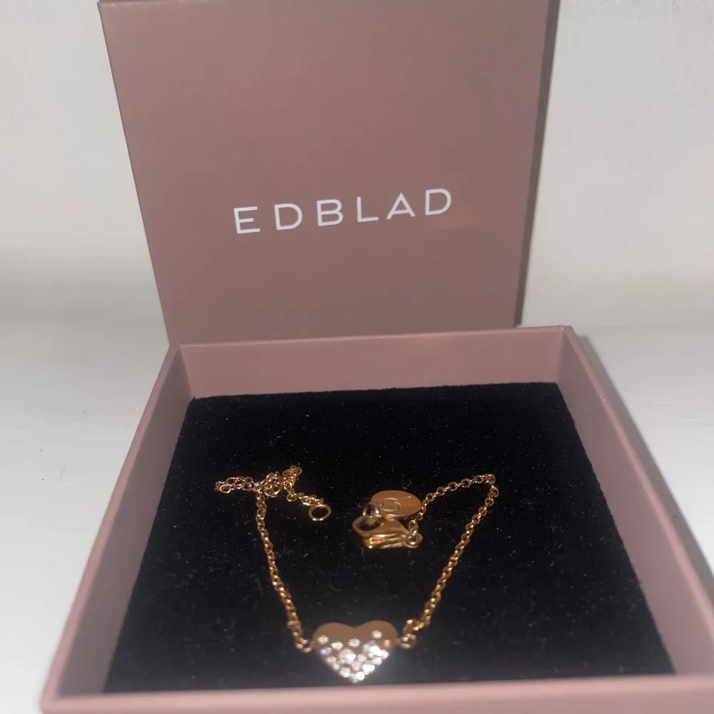 Elegant guldarmband från Edblad med ett hjärta dekorerat med små glittrande stenar. Perfekt för att ge en touch av glamour till din outfit. Armbandet har en justerbar kedja och kommer i en fin presentask.. Asusteet.