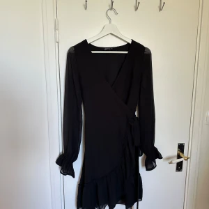 Svart omlottklänning från Gina Tricot - Elegant svart omlottklänning från Gina Tricot med långa ärmar och volangdetaljer. Perfekt för en stilren look med knytband i midjan och lätt transparenta ärmar.