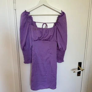 Lila klänning med puffärmar - Charmig lila klänning med puffiga trekvartsärmar och smockdetaljer vid ärmsluten. Klänningen har en öppen rygg med knytband för en extra touch. Perfekt för vår och sommar.