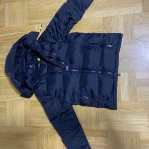 Svart dunjacka från Polo Ralph Lauren - Snygg svart dunjacka från Polo Ralph Lauren med huva och dragkedja. Jackan har en klassisk pufferdesign och är perfekt för kyliga dagar. Den har långa ärmar och praktiska fickor med dragkedja.