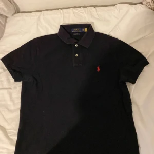 Svart pikétröja från Ralph Lauren - Snygg svart pikétröja från Ralph Lauren med klassisk krage och två knappar. Den har en röd broderad logotyp på bröstet och är i en slim fit passform. Perfekt för en stilren look.