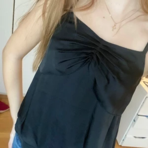 Svart linnetopp från JC - Snygg svart linnetopp från JC med smala axelband och rynkdetalj framtill. Perfekt för en stilren look. Tillverkad i ett mjukt och följsamt material.