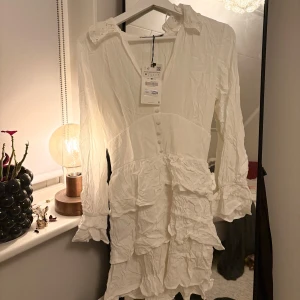 Vit klänning från Zara - Säljer en vit klänning från Zara med volanger och knappar framtill. Klänningen har långa ärmar och en v-ringad design. Helt oanvänd, lapp kvar