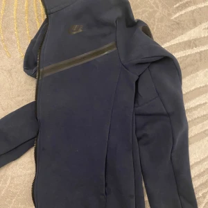 Mörkblå nike tech  - Säljer en mörkblå jacka från Nike med dragkedja och diskret logga på bröstet. Jackan har en stilren design med svarta detaljer längs dragkedjan. Perfekt för en sportig look. Men har få skador kolla 2 sista bilderna. Pris kan diskuteras 