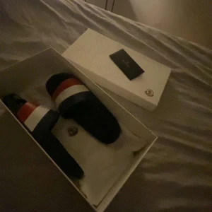  tofflor från Moncler - Snygga svarta sandaler från Moncler med röd, vit och blå rand över foten. Perfekta för en avslappnad stil med en touch av lyx. Kommer i originalförpackning. Passar 39 pris ej hugget!