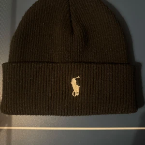 Svart ribbad mössa från Ralph Lauren - Snygg svart ribbad mössa från Ralph Lauren med broderad logga framtill. Perfekt för kyliga dagar och en stilren look.