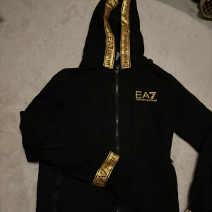 Svart hoodie från EA7 Emporio Armani - Snygg svart hoodie från EA7 Emporio Armani med gulddetaljer. Den har en dragkedja framtill och märkeslogga på bröstet. Perfekt för en stilren och bekväm look.