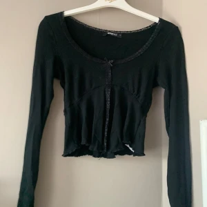 Svart croppad kofta från Gina Tricot - Säljer en svart croppad kofta från Gina Tricot med spetsdetaljer längs halsringningen och slits framtill. 