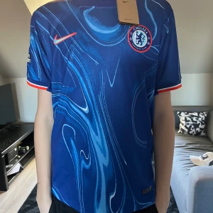 Blå Chelsea fotbollströja från Nike - Säljer en blå Chelsea fotbollströja från Nike med ett unikt mönster och klubbens emblem på bröstet. Tröjan har korta ärmar med röda detaljer vid ärmsluten. Perfekt för alla Chelsea-fans! 💙