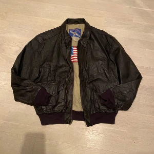 Brun  skinnjacka från Airborne - Säljer en mörkbrun skinnjacka från Airborne med dragkedja och ribbade muddar. Jackan har en cool amerikansk flagga på insidan och är tillverkad i äkta läder.