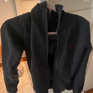Svart hoodie från Ralph Lauren - Snygg svart hoodie från Ralph Lauren med dragkedja och en liten röd logga på bröstet. Perfekt för en avslappnad stil.