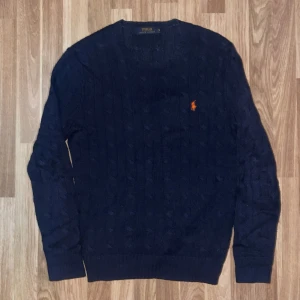 Mörkblå stickad tröja från Polo Ralph Lauren - Säljer en snygg mörkblå stickad tröja från Polo Ralph Lauren med det klassiska broderade logotypen i röd på bröstet. Tröjan har långa ärmar och en ribbad struktur som ger en stilren look. Perfekt för kyligare dagar.