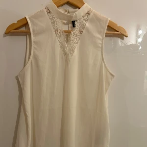 Vit ärmlös topp från Vero Moda - Säljer en stilren vit ärmlös topp från Vero Moda. Toppen har en hög hals med spetsdetaljer framtill och en liten öppning i nacken. Perfekt för en elegant look.
