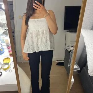 Vit ärmlös topp med spetsdetaljer - Säljer en söt vit ärmlös topp med spetsdetaljer och smala axelband. Toppen har en luftig passform och ett vackert broderat mönster. Perfekt för varma sommardagar!