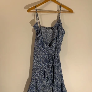 Blå blommig klänning med volangdetaljer - Charmig blå klänning med vita blommor och volangdetaljer. Den har smala axelband och en knytning i midjan för en fin passform. Perfekt för somriga dagar.