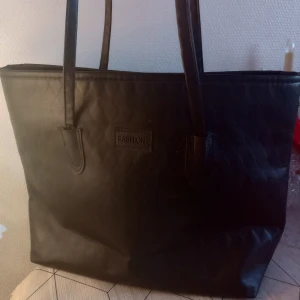 Svart handväska - Säljer denna snygga svarta handväskan. Väskan har två långa handtag och en rymlig insida, perfekt för att bära med sig allt man behöver. Märkt med 'Fashion' på framsidan.