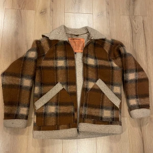 Tjock foderjacka från Carlings - Säljer en brun rutig jacka från Carlings med mysigt teddyfoder. Jackan har dragkedja och två snedställda fickor framtill. Perfekt för kyliga dagar med sin varma och stiliga design.