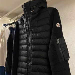 Moncler Cardigan - Säljer en snygg svart jacka från Moncler med quiltad design och stickade ärmar. Jackan har en dragkedja framtill och en praktisk ficka med dragkedja på ärmen. Perfekt för kyligare dagar med sin stilrena look och bekväma passform. Strl L Äkta hör av dig om du har några frågar 