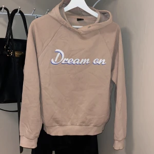 Beige hoodie från Gina Tricot - Säljer en beige hoodie från Gina Tricot med texten 'Dream on' i vitt och blått på framsidan. Tröjan har en mysig huva och långa ärmar, perfekt för en avslappnad stil.