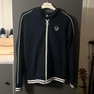 Marinblå huvtröja från Sergio Tacchini - Snygg marinblå huvtröja från Sergio Tacchini. Tröjan har långa ärmar och en bekväm passform. Perfekt för en avslappnad stil.