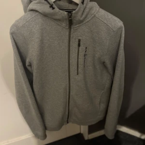Grå huvtröja från Sail Racing - Snygg grå huvtröja från Sail Racing med dragkedja och huva. Perfekt för en avslappnad stil. Tröjan har en bröstficka med dragkedja och är tillverkad i ett mjukt material för extra komfort. Är i riktigt bra gick och säljs pga att den är för liten. 