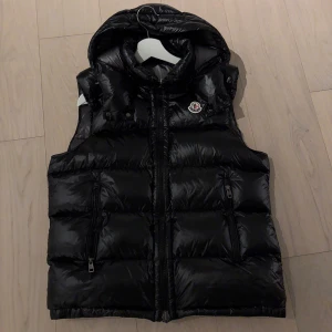 Moncler väst svart - Snygg svart dunväst från Moncler med dragkedja och huva. Perfekt för kyliga dagar när du vill hålla stilen. Västen har två praktiska fickor med dragkedja och Monclers ikoniska logga på bröstet. Knappt använd, köpt är köpt