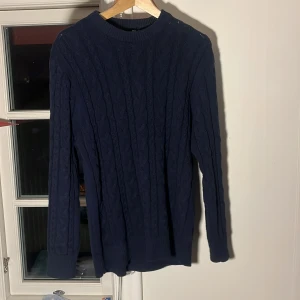 Mörkblå stickad tröja - Snygg mörkblå stickad tröja med kabelstickat mönster. Perfekt för kyliga dagar med sin klassiska design och långa ärmar. Passar bra till både jeans och chinos.