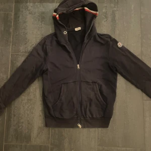 Moncler tröja - En klassisk Moncler-tröja i storlek large, men med en passform som känns som medium. Tröjan är tillverkad i mjuk och bekväm bomullsblandning, perfekt för vardagsbruk. Designen är stilren med Monclers ikoniska logga placerad på sidan. Observera att tröjan har en reva vid armbågen och solblekta partier som ger den en använd look. Ett plagg för dig som uppskattar både kvalitet och karaktär.