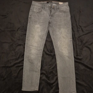 Grå jeansbyxor från Jack & Jones - Snygga grå jeansbyxor från Jack & Jones med en klassisk femficksdesign. Byxorna har en dragkedja och knappstängning. Perfekta för en avslappnad stil.