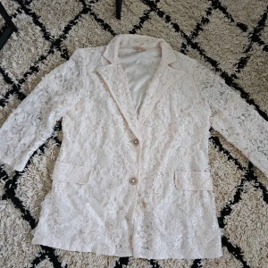 Champagnefärgad spetskavaj  med pärlknappar - Elegant  spetskavaj med vackra pärlknappar och trekvartsärmar. Perfekt för en stilfull look med en feminin touch. Kavajen har en klassisk krage och två fickor framtill. Från Buch Copenhagen. 