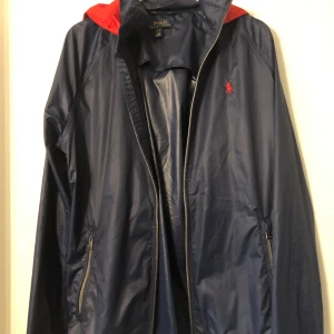 Blå vindjacka från Polo Ralph Lauren - Snygg blå vindjacka från Polo Ralph Lauren med röd insida och broderad logga på bröstet. Jackan har dragkedja och praktiska fickor med dragkedjor. Perfekt för blåsiga dagar.