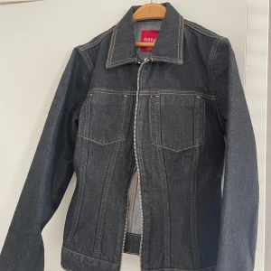 Svart jeansjacka från MNG - Snygg svart jeansjacka från MNG med dragkedja framtill och klassisk krage. Jackan har långa ärmar och två bröstfickor med knappdetaljer. Perfekt för en stilren look.