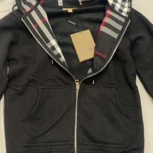 Svart huvtröja med dragkedja - Snygg svart huvtröja från Burberry med dragkedja och klassiskt rutigt mönster i huvan. Perfekt för en stilren look med en touch av lyx. Tröjan har praktiska fickor framtill och en bekväm passform.