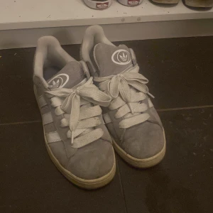 Adidas Campus 00s Gray White - Snygga grå Adidas sneakers med vita detaljer och breda snören. Skorna har den klassiska tre-randiga designen på sidorna och Adidas-loggan på plösen och hälen. Perfekta för en avslappnad stil.
