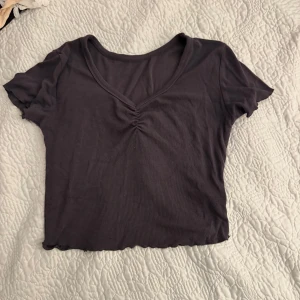 Mörkgrå ribbad topp - Säljer en mörkgrå ribbad topp med v-ringning och rynkdetalj framtill. Toppen är kortärmad och har en croppad passform, perfekt för en avslappnad stil.