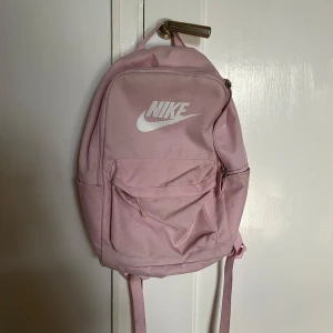 Rosa ryggsäck från Nike - Säljer en snygg rosa ryggsäck från Nike med vit logga på framsidan. Ryggsäcken har flera fack med dragkedja och justerbara axelremmar. Perfekt för skolan eller träningen!
