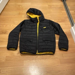Svart pufferjacka från Barbour - Snygg svart pufferjacka från Barbour med gul insida. Jackan har en huva och dragkedja framtill samt tre fickor med knappar. Perfekt för kyligare dagar med sin quiltade design och bekväma passform. Jag är 168 och den passar bra men är bara lite kort i armarna💕skriv till mig innan köp